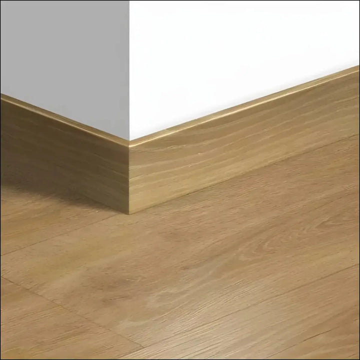 Gold-finished quickstep majestic skirting boards 58mm with subtle wood grain on light gray wall and hardwood floor corner